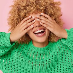Happy young woman with curly hair covers eyes awaits for surprise smiles broadly shows white teeth dressed in casual knitted green jumper feels glad isolated over pink background. Blind concept